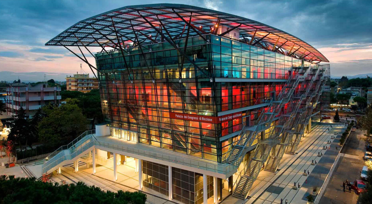 Exterior view of the Palazzo dei Congressi di Riccione, a modern glass conference center with a curved metal roof and long external staircases at dusk, illuminated inside and outside.